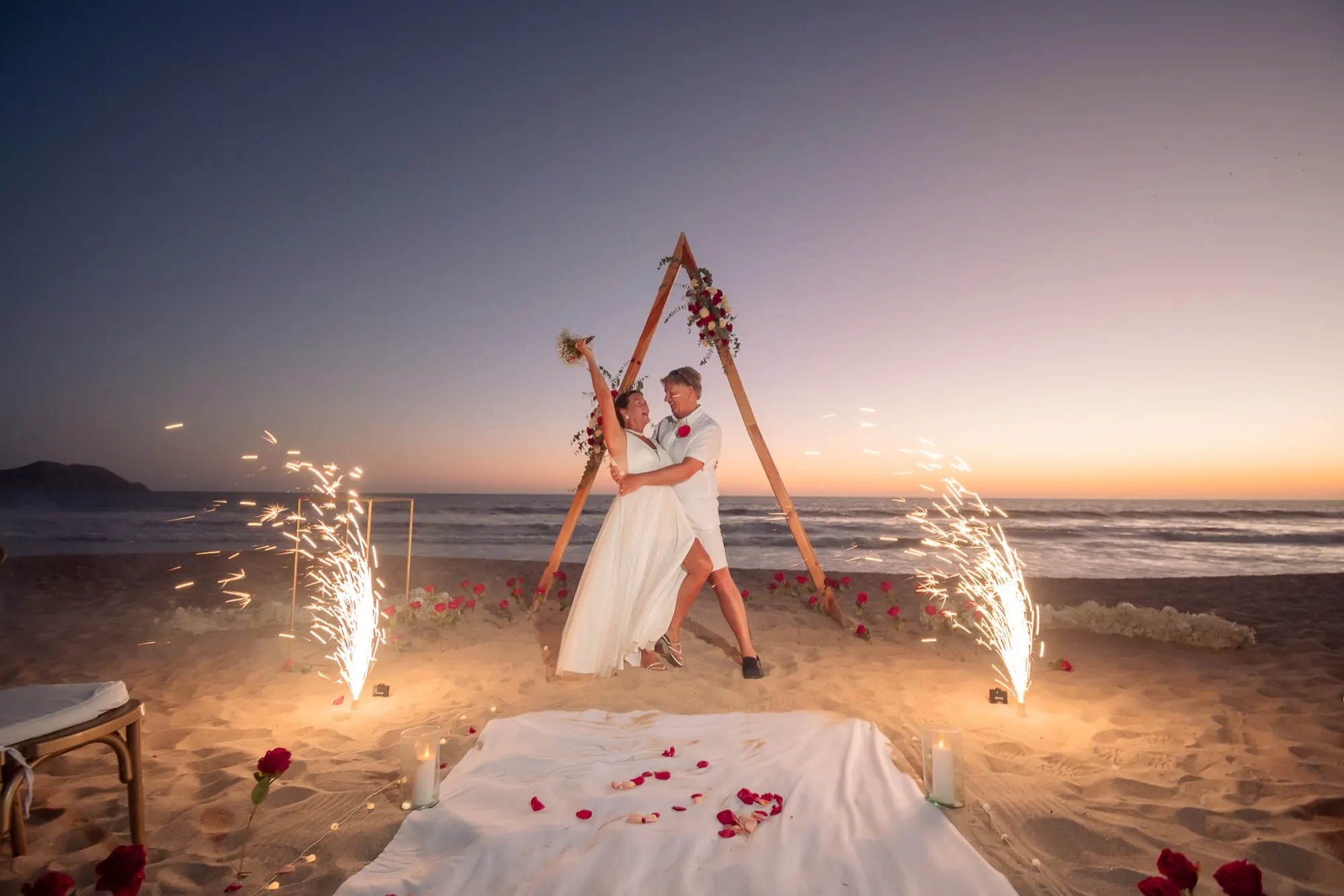 Pareja bailando bajo arco floral en la playa con chispas al atardecer